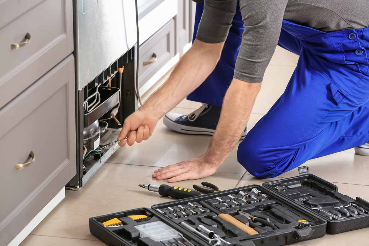 Repair man kneeling to tinker with electrical wires on bottom of refrigerator